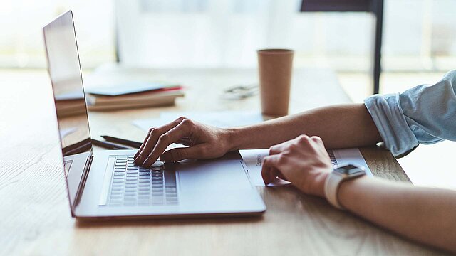 Schreibtisch mit einem geöffneten Laptop, Hand auf der Tastatur
