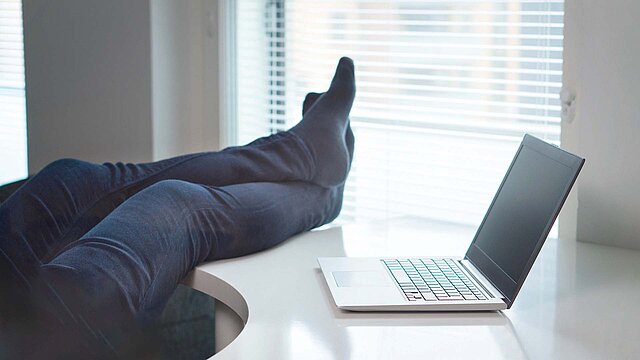 ein geöffneter Laptop stehend auf einem weißen Schreibtisch, verschränkte Beine einer Person liegen quer über dem Schreibtisch, im Hintergrund Fenster mit Jalousie