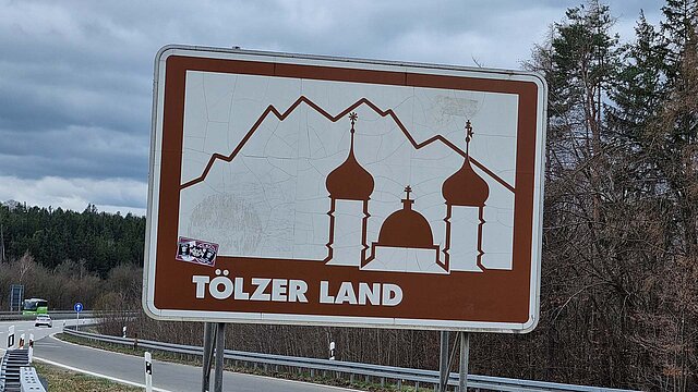 Touristisches Hinweisschild in braun/weißer Farbe mit Aufschrift "Tölzer Land“ an der Autobahn stehend